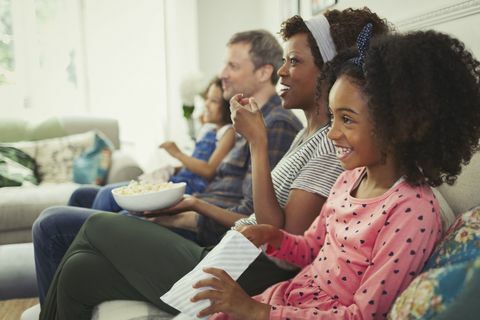 युवा बहु-जातीय परिवार फिल्म देखते हैं और सोफे पर पॉपकॉर्न खाते हैं